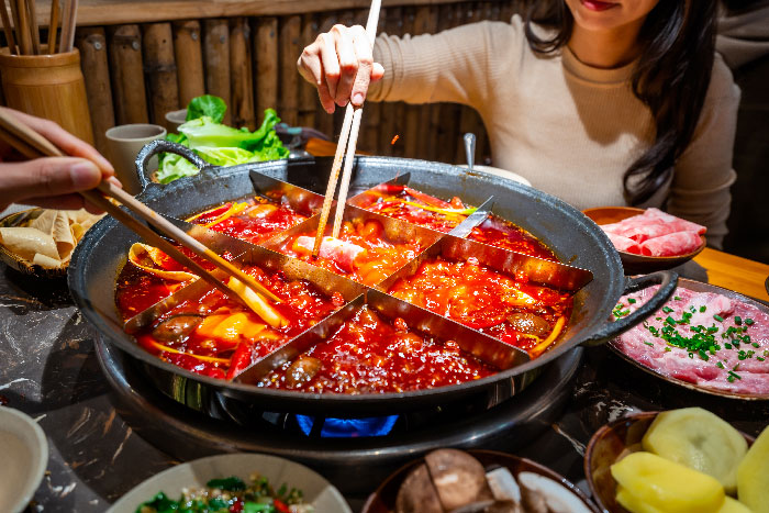 Primer plano de un hot pot tradicional de Chongqing dividido en cuadrículas con caldo picante rojo burbujeando, mientras varias personas usan palillos para cocinar.