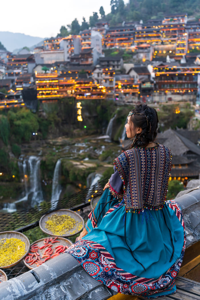 Mujer vestida con ropa tradicional sentada en un tejado, contemplando una ciudad antigua iluminada en la montaña y cascadas al fondo.