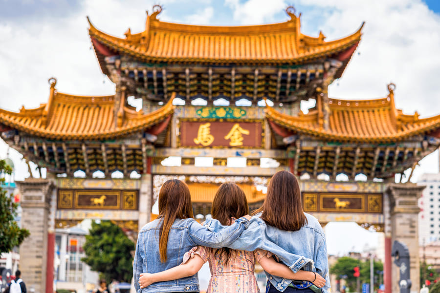Tres mujeres de espaldas abrazadas contemplando una gran puerta tradicional china con tejados dorados y ornamentos tallados.
