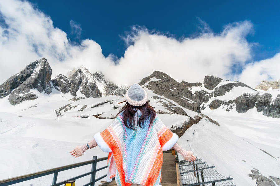 Mujer de espaldas con los brazos abiertos en un sendero de madera, contemplando las montañas nevadas de la Montaña de Nieve del Dragón de Jade bajo un cielo azul con nubes.
